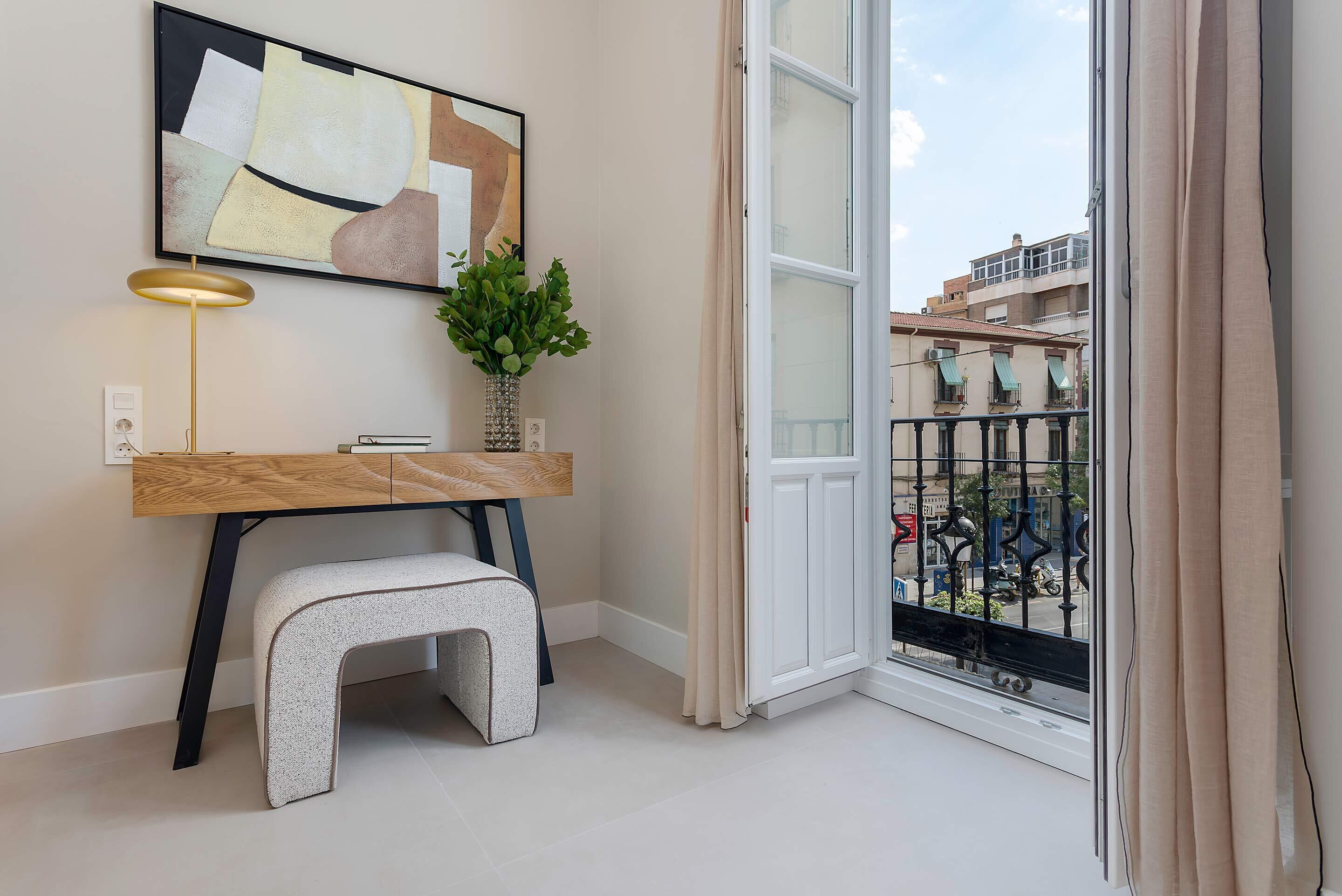 A modern living room with a wooden desk, a small stool, a potted plant, and a large window with a view of the city.