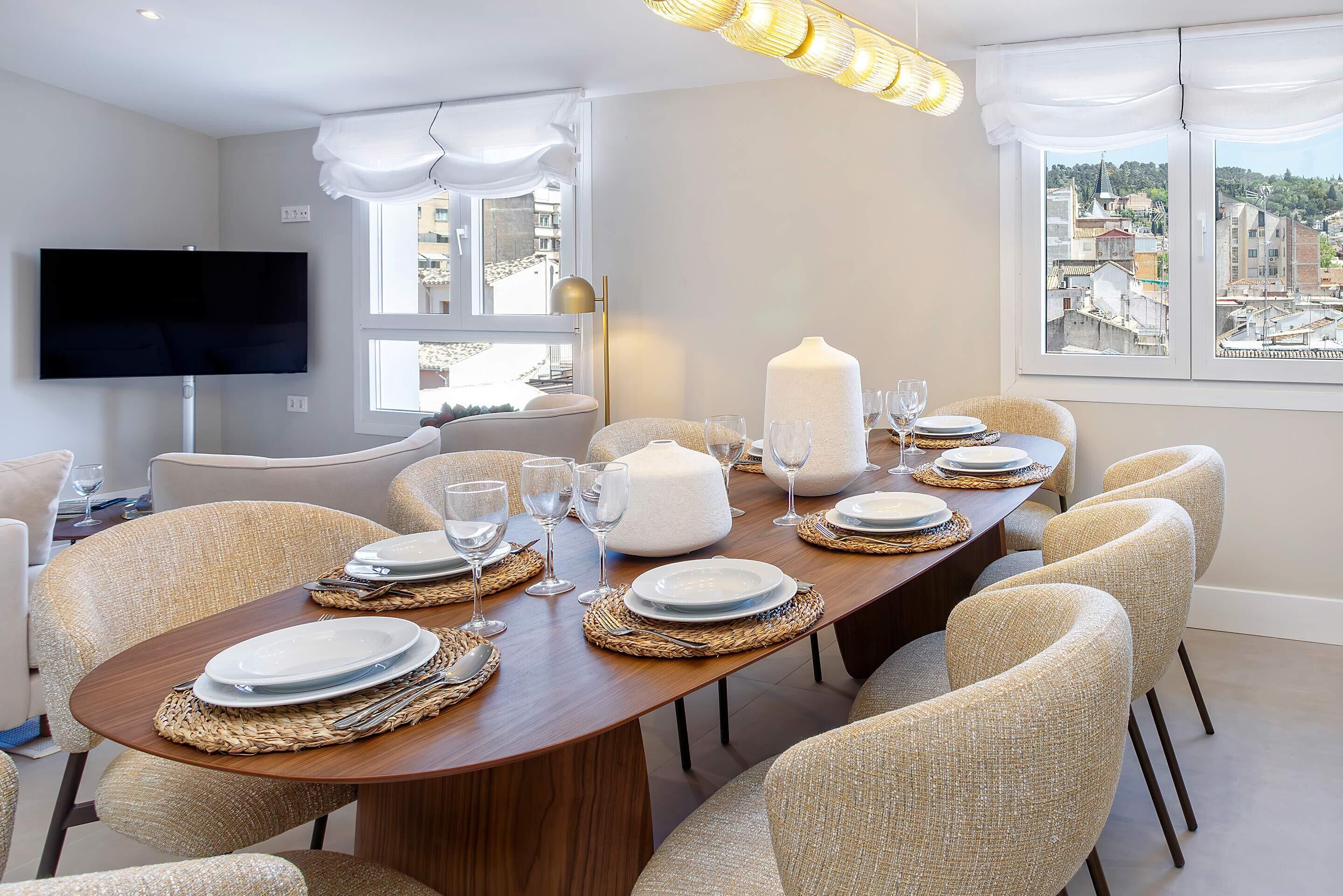 A dining area with a long wooden table set for six, surrounded by beige upholstered chairs.