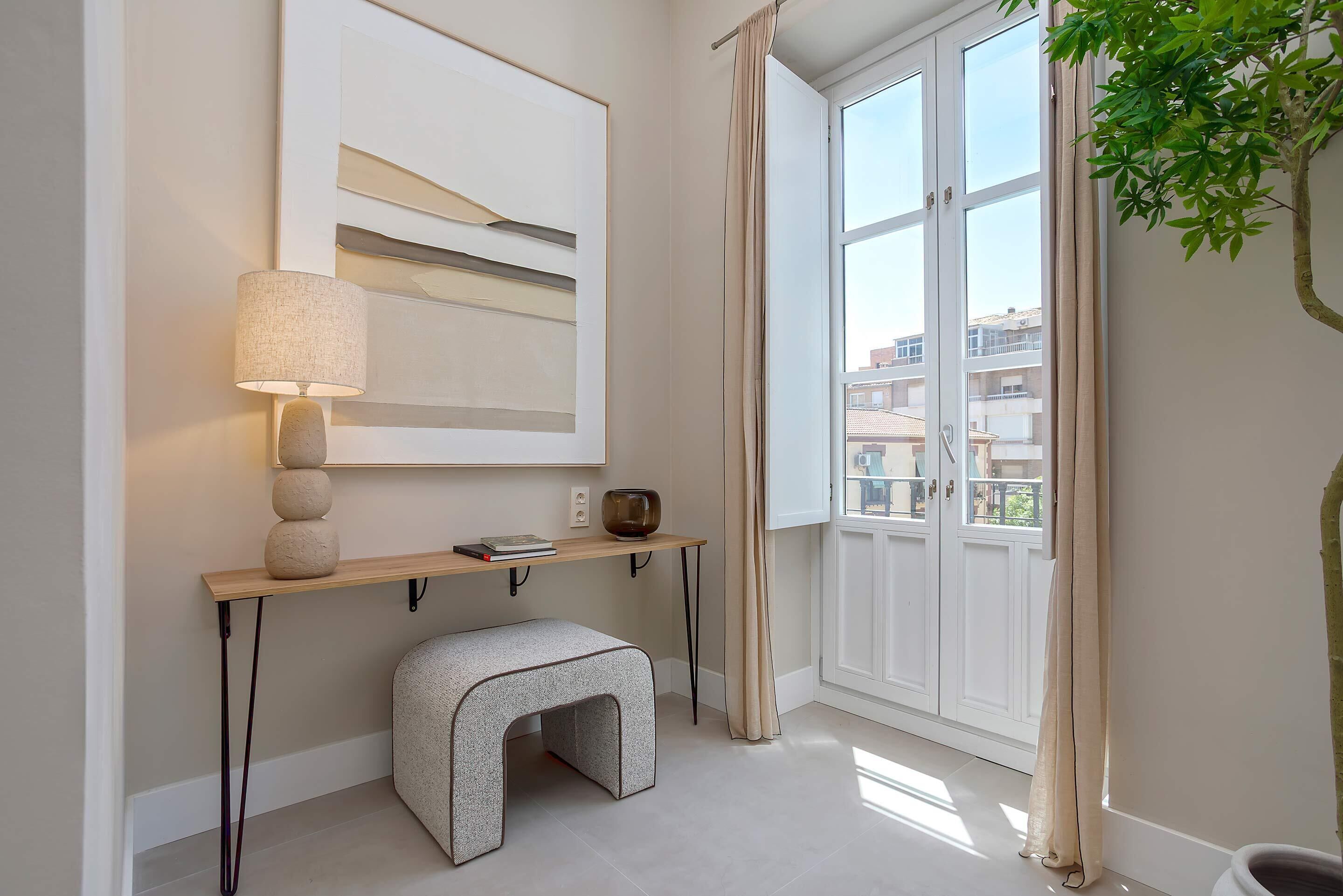 A modern room with a large window, a wall-mounted lamp, a bench, and a shelf with a book and a bowl.
