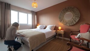 Ein Hotelzimmer mit Bett, Nachttischlampen, einem runden Spiegel, einem Holzstuhl und einem Fenster mit Vorhängen.