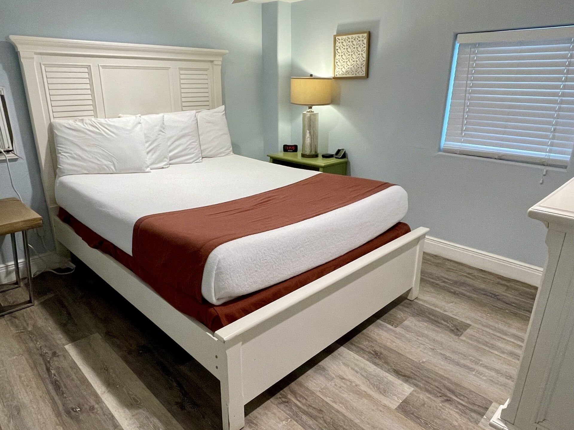 A bed with white bedding and a brown accent, a bedside table with a lamp, a wall-mounted decorative piece, and a window with blinds.