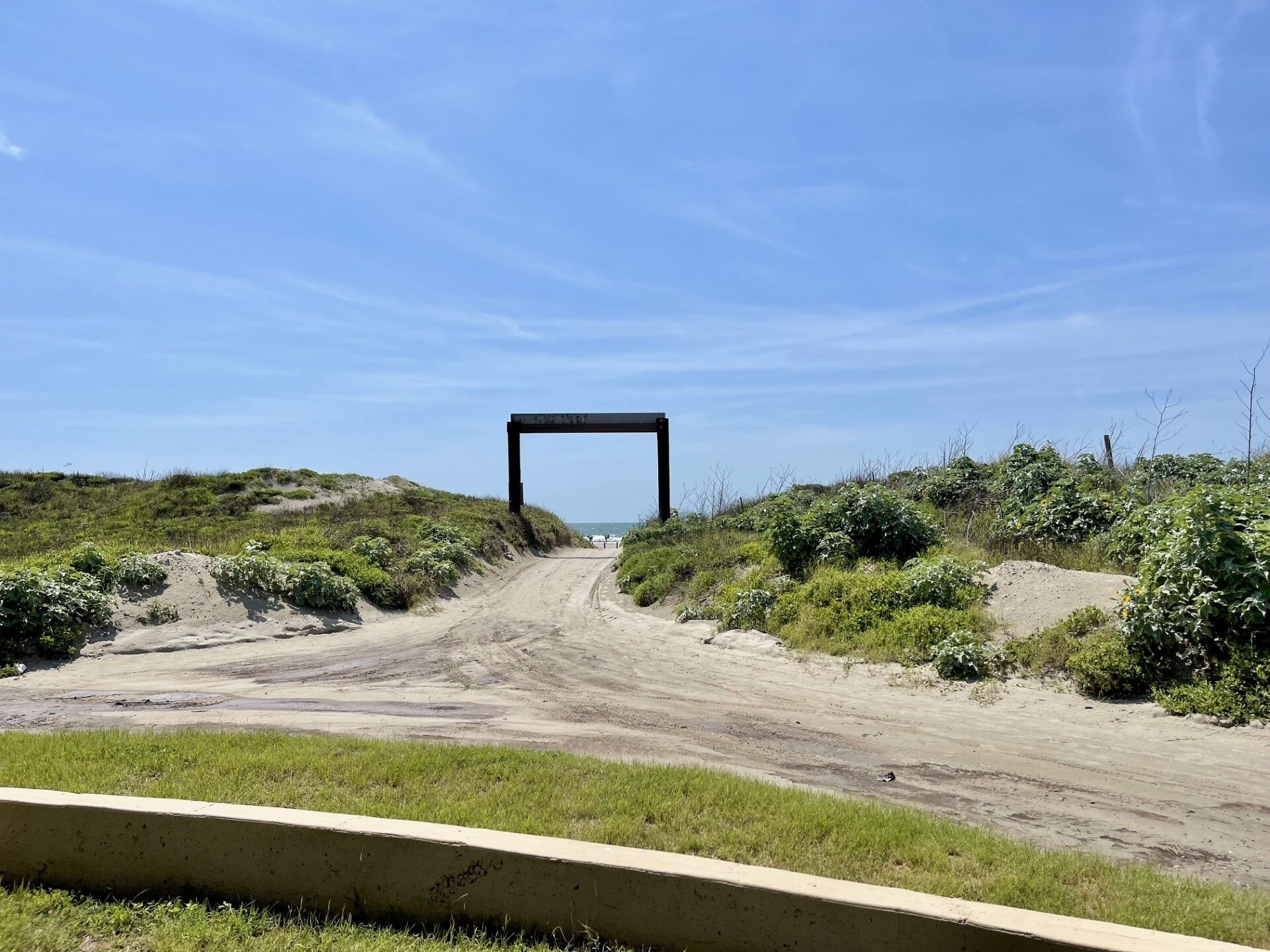 A dirt road leading to a black metal structure, surrounded by greenery and a clear sky.