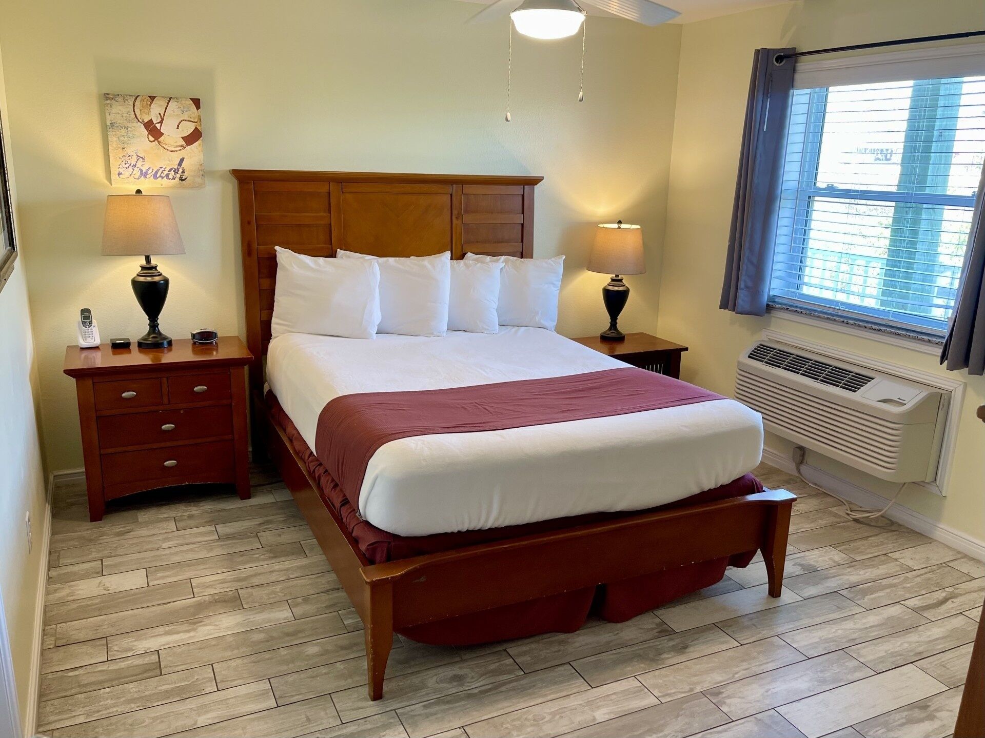 A hotel room with a wooden bed, two bedside tables, a wall-mounted air conditioner, and a window with blinds.