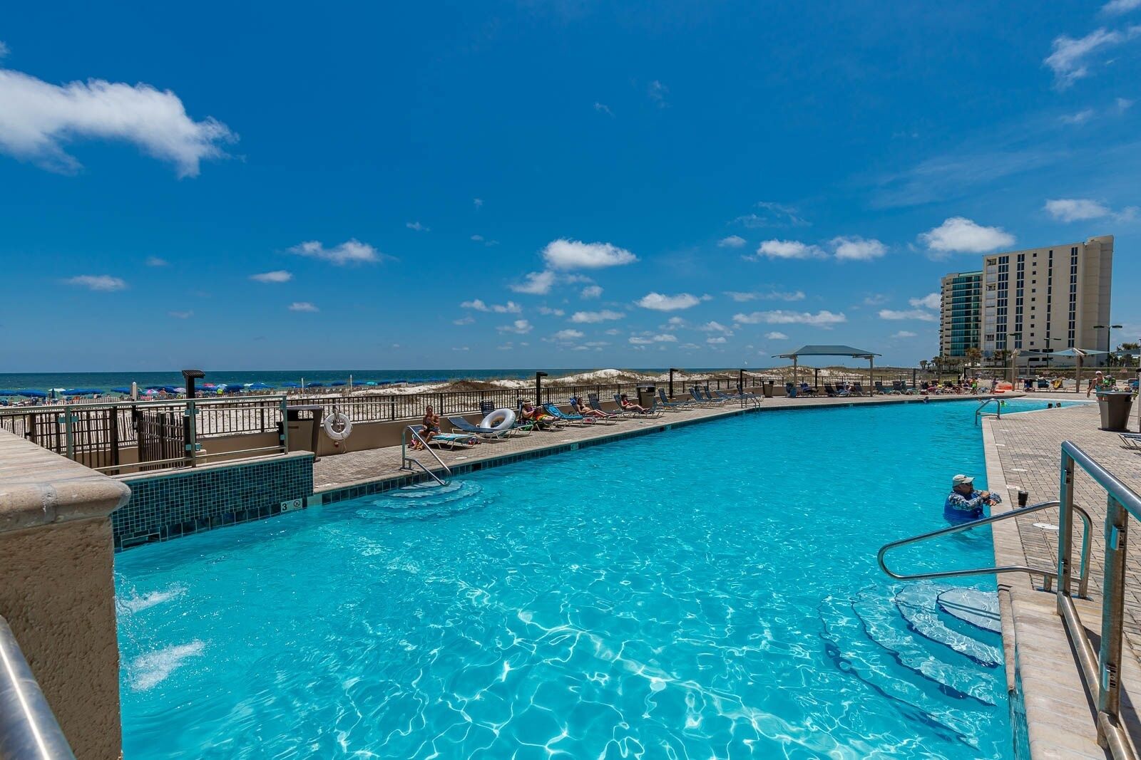 A swimming pool with clear blue water, surrounded by lounge chairs and umbrellas, with a beach and ocean in the background.