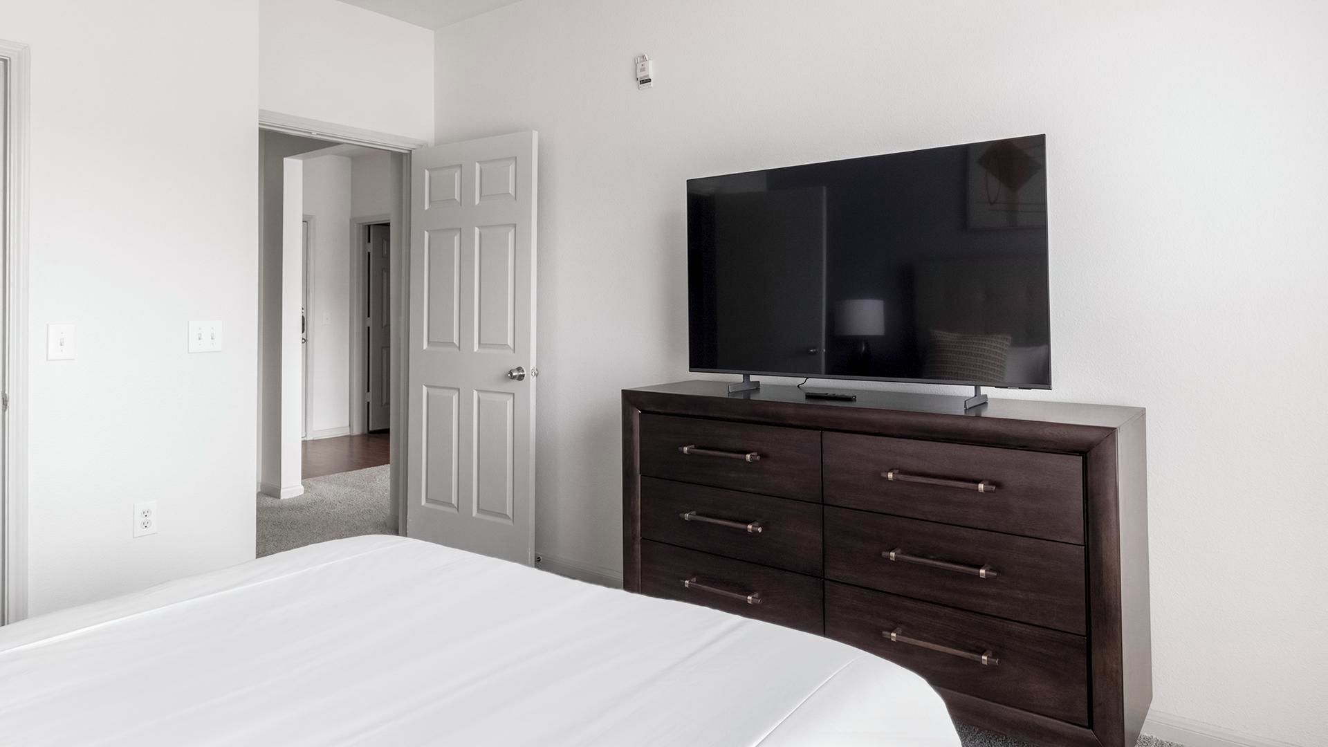 A bedroom with a bed, a dark wooden dresser, a television mounted above it, and a white door.