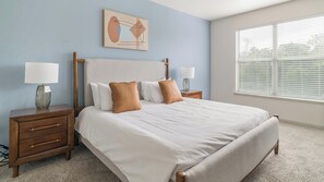 A bedroom with a large bed, two bedside tables, a nightstand, a window with blinds, and a wall-mounted artwork.