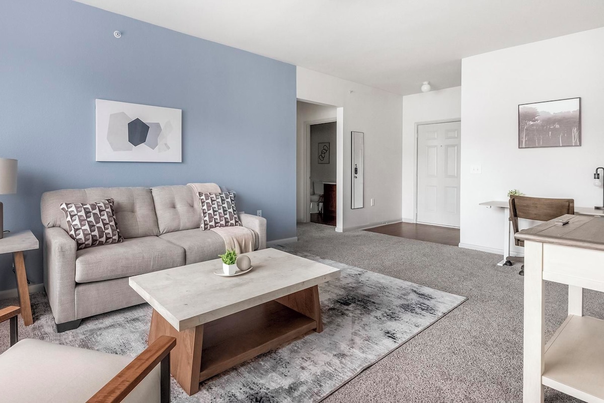 A modern living room with a sofa, coffee table, and a lamp. There is a door leading to another room.