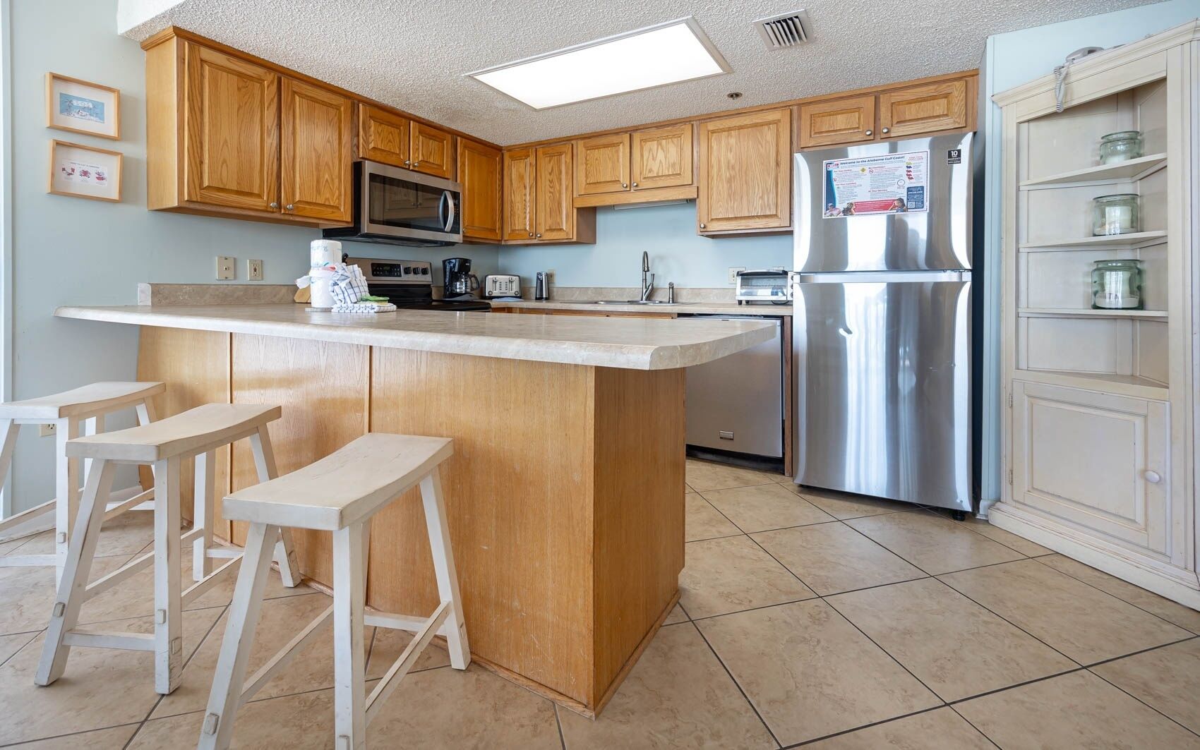 A kitchen with wooden cabinets, a stainless steel refrigerator, a microwave, and a bar stool area.