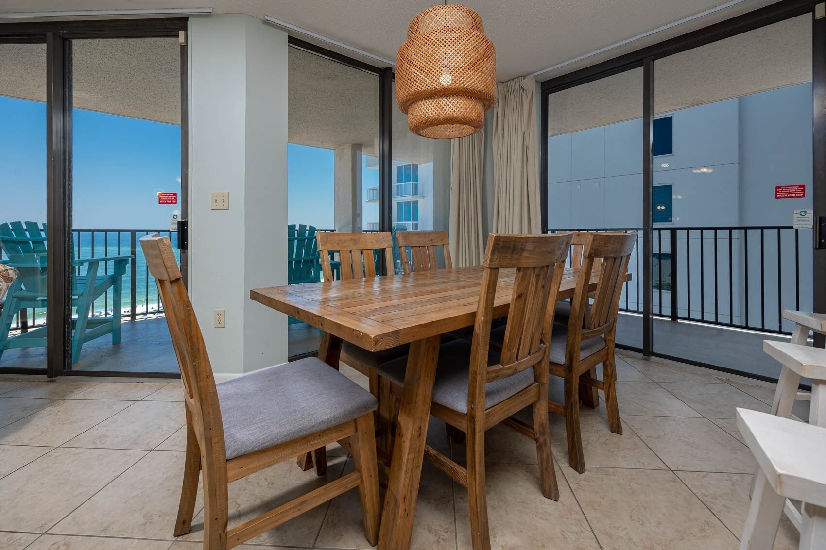 A wooden dining table with six chairs, a wicker pendant light, and a view of the ocean through a sliding glass door.