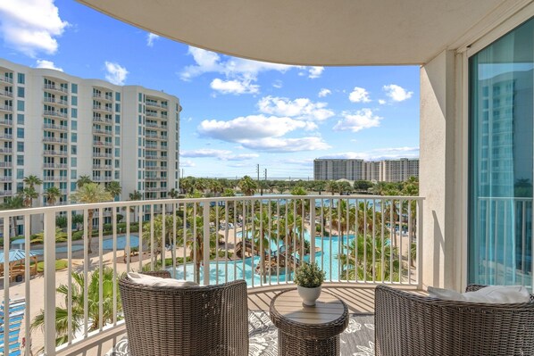 A balcony with wicker furniture, a view of a swimming pool, and high-rise buildings in the background. - Gulfview Retreat At The Palms 2 Bedroom Condo (Destin)