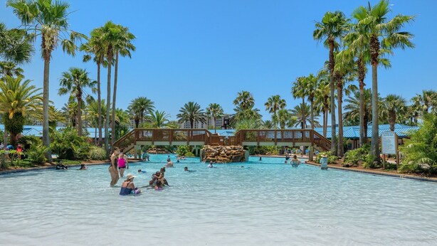 A large outdoor swimming pool surrounded by palm trees and a wooden deck. - Gulfview Retreat At The Palms 2 Bedroom Condo (Destin)