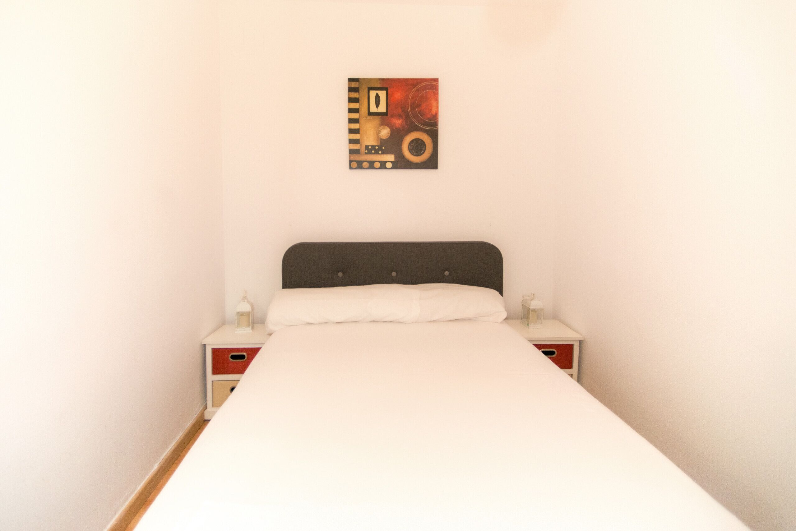 Un lit avec un matelas blanc et une tête de lit sombre, flanqué de deux tables de chevet dotées de tiroirs rouges et d’une lampe blanche sur chacune.