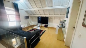 Ein modernes Wohnzimmer mit Fernseher, zwei weißen Sesseln, einem Glastisch und einer kleinen Küchenzeile inklusive Kühlschrank.