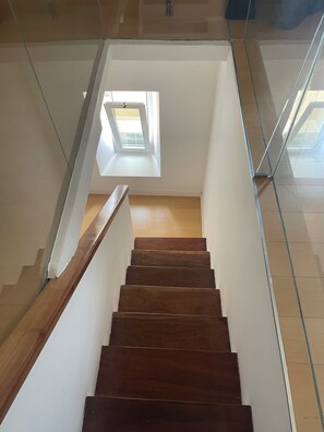 A wooden staircase with a glass railing, leading to a room with a skylight.