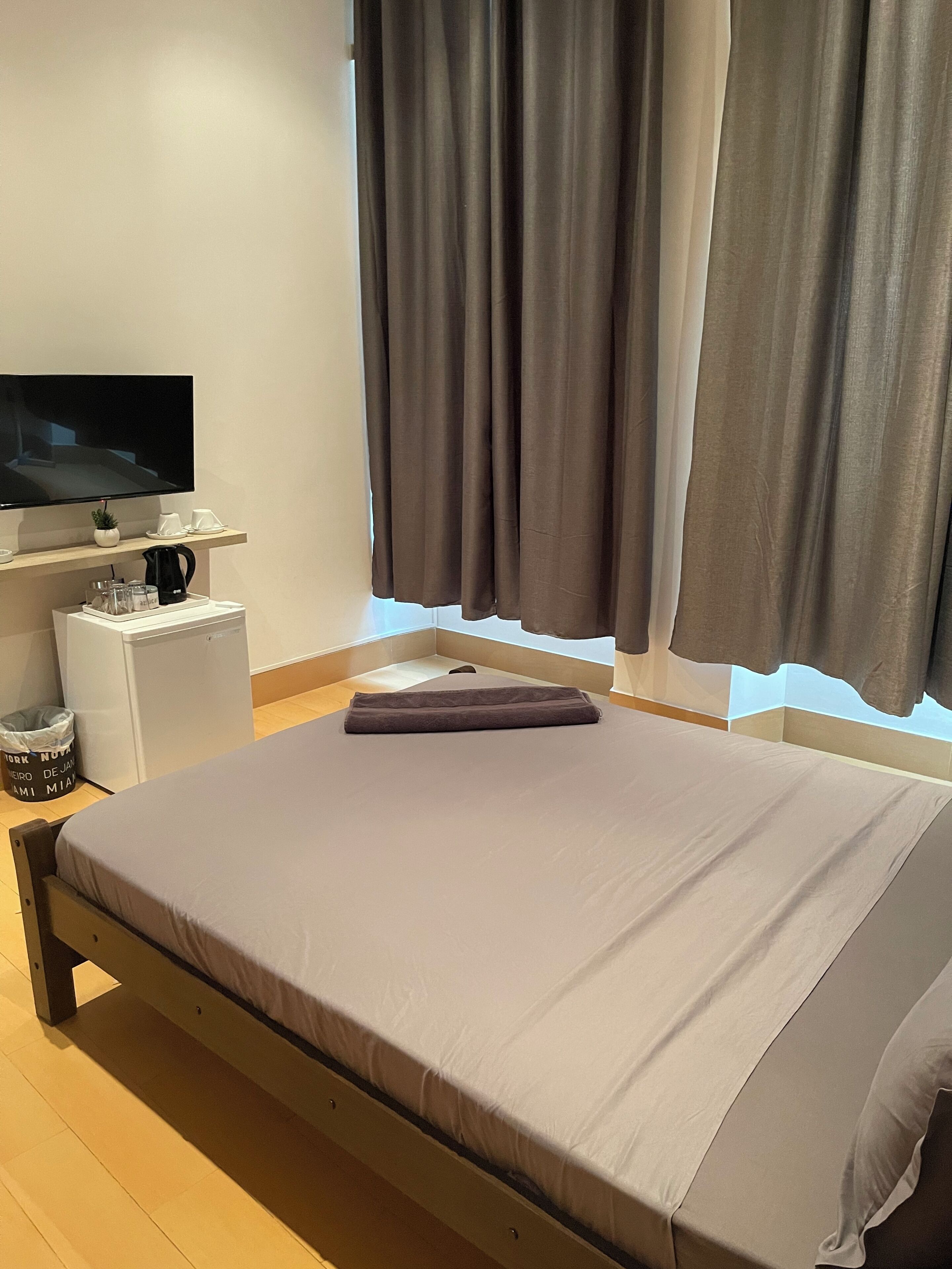 Una cama bien hecha con una manta doblada, un televisor de pantalla plana montado encima, una mesita blanca con una cafetera y una ventana con cortinas.