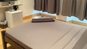 Ein ordentlich bezogenes Bett mit gefalteter Decke, ein flachbildfernseher darüber angebracht, ein kleiner weißer Tisch mit Kaffeemaschine und ein Fenster mit Vorhängen.