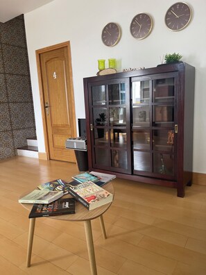 A room with a wooden door, a glass-top table with books, a dark wooden cabinet with glass doors, and three wall clocks.