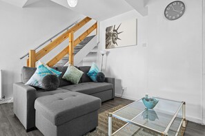 Ein modernes Wohnzimmer mit einem grauen Ecksofa, einem Glastisch, einer Treppe mit Holzwandleisten und einer Wanduhr.