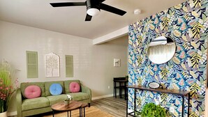 Un salon avec un canapé vert, un ventilateur de plafond, un mur à motifs et une petite table surmontée d’un miroir.