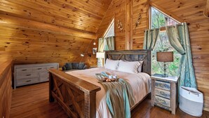Ein Schlafzimmer einer Holzhütte mit Bett, Nachttisch, Lampe und einem Fenster mit Vorhängen.