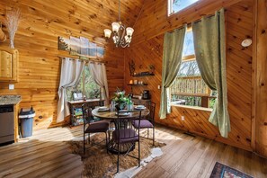 Interior de una cabaña de madera con una mesa de comedor, sillas, una araña y vistas al exterior.