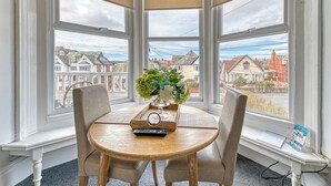 Et karnapvindue med tre ruder, et rundt træbord med to stole, en vase med blomster, en fjernbetjening og en vinduesplads.