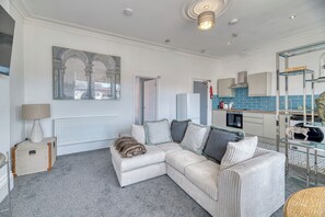 Ein modernes Wohnzimmer mit einem Ecksofa, einem Heizkörper, einer Küchenzeile mit blauer Spritzschutz und einem großen gerahmten Kunstwerk an der Wand.