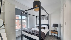 En seng med baldakin, et sengebord med lampe, og billeder i rammer på væggen.