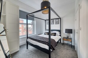 Ein Bett mit Baldachin, ein Nachttisch mit Lampe sowie gerahmte Bilder an der Wand.