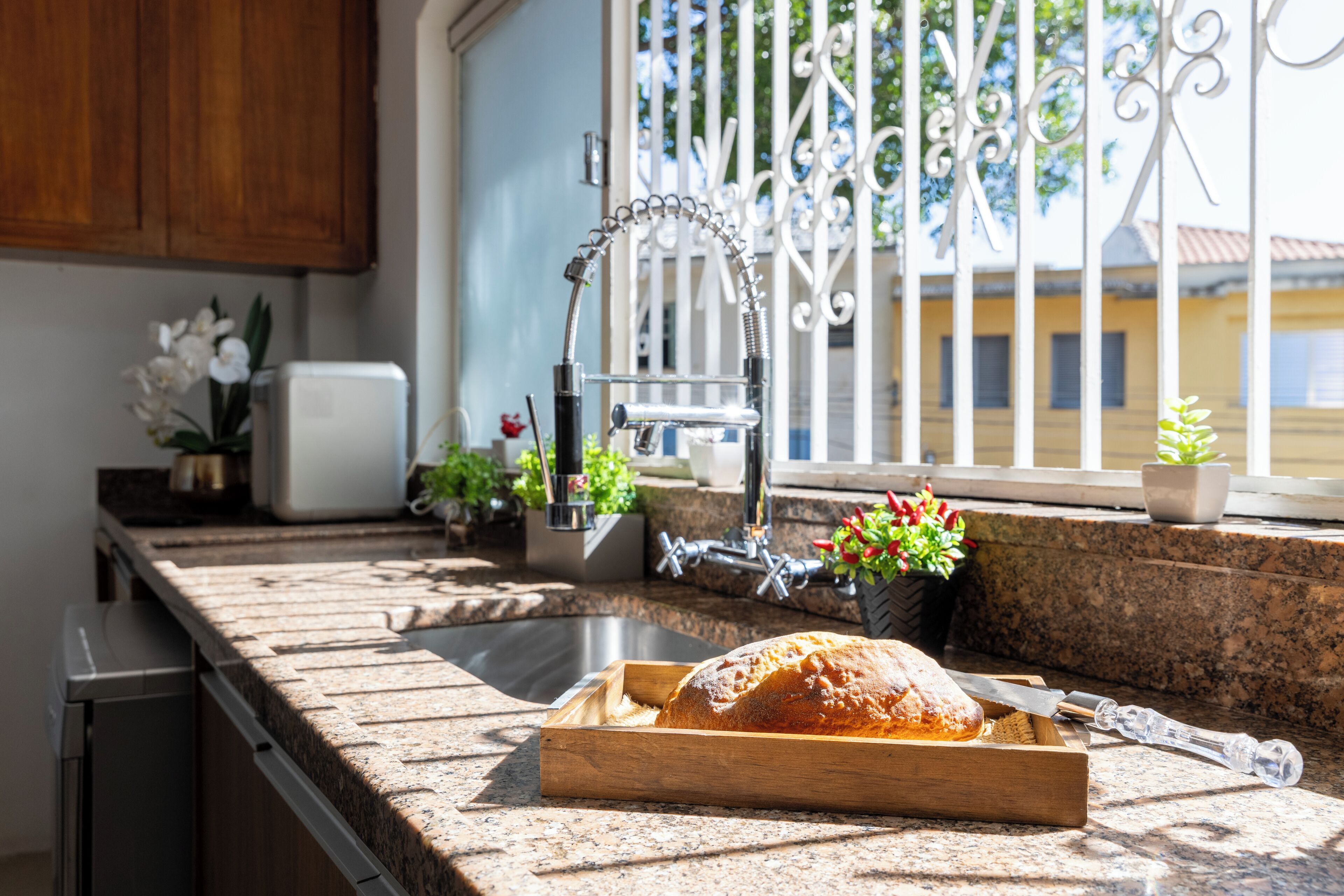 A kitchen with a granite countertop, a bread loaf on a wooden cutting board, a stainless steel sink, and a window with a metal gate.