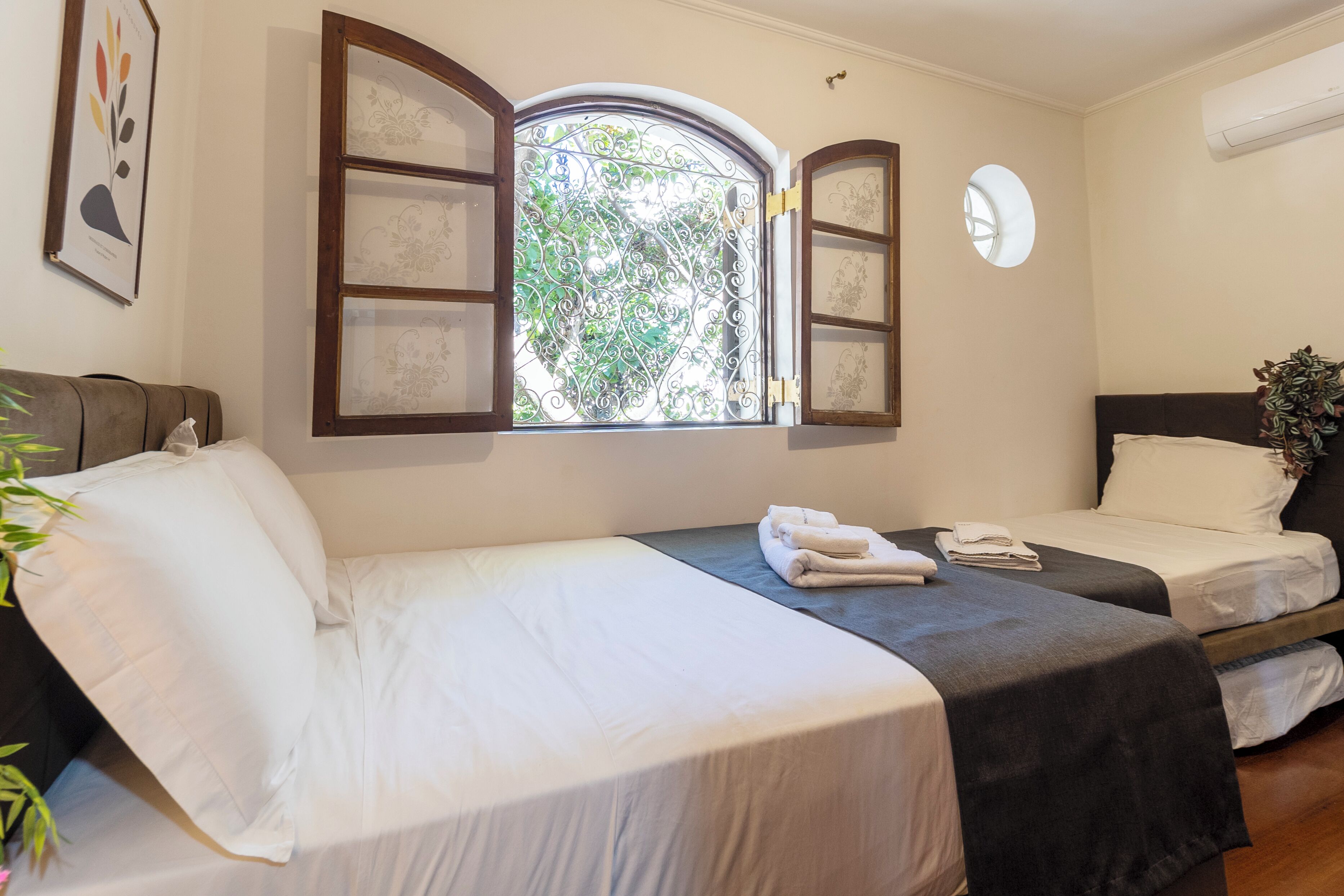 A hotel room with two beds, a window with open shutters, a wall-mounted air conditioning unit, and a framed picture.