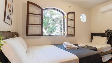 A hotel room with two beds, a window with open shutters, a wall-mounted air conditioning unit, and a framed picture.