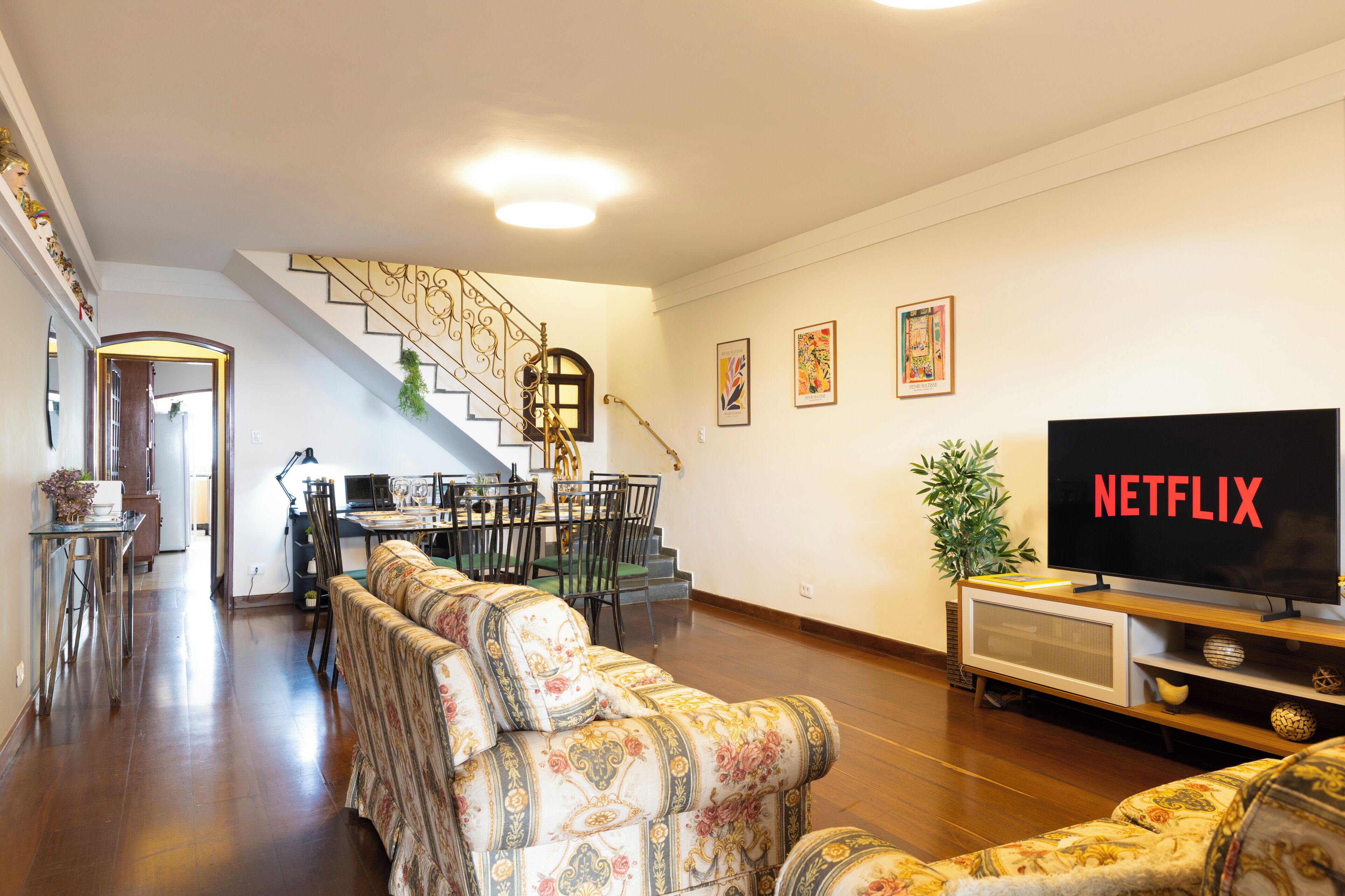 A living room with a sofa, a dining area with chairs and a table, a staircase, and a television displaying 'NETFLIX'.