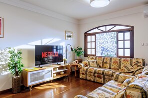A living room with a flat-screen TV displaying 'NETFLIX,' a sofa, a coffee table, a potted plant, and a window with decorative glass. - Casa de 4 quartos com AC no Ipiranga (São Paulo)