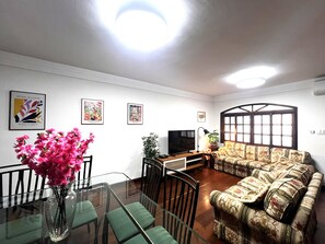 A living room with a glass dining table, green chairs, a sofa, a TV, and wall art. - Casa de 4 quartos com AC no Ipiranga (São Paulo)