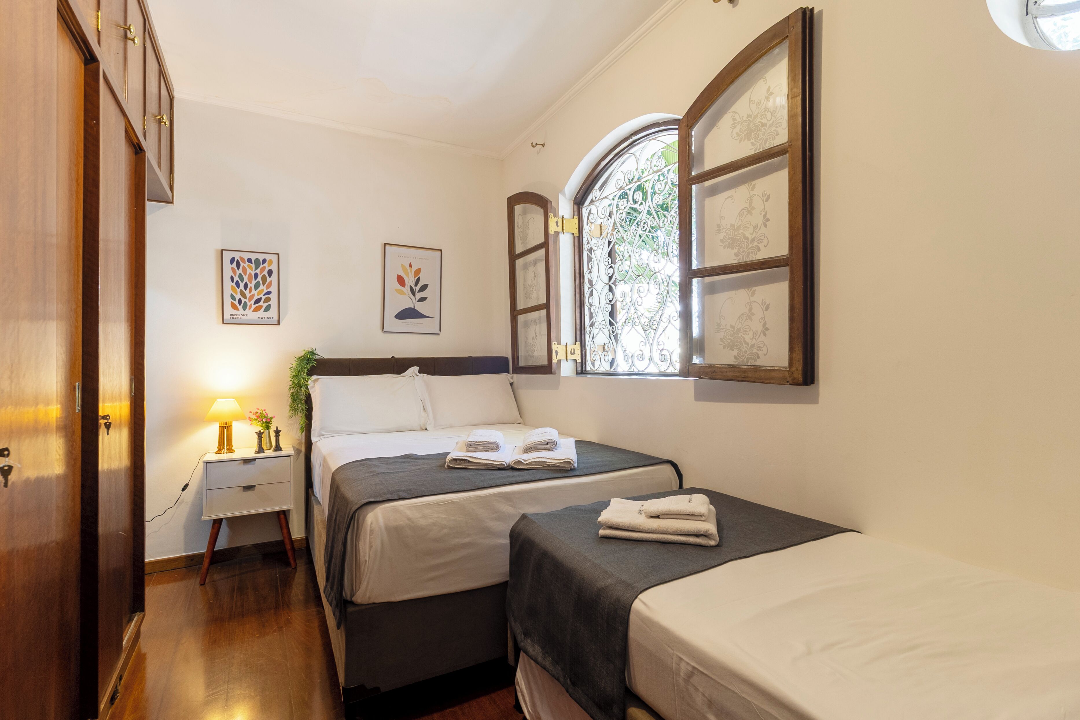 Une chambre avec deux lits, une table de chevet avec une lampe, une fenêtre ornée de vitraux et des œuvres d’art murales.