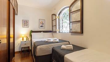 Quarto com duas camas, mesa de cabeceira com abajur, janela com detalhe decorativo e arte na parede.