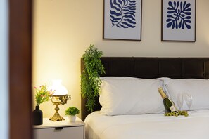 A neatly made bed with white linens, a bedside table with a lamp and a small plant, and a bottle of champagne with glasses on the bed. - Casa de 4 quartos com AC no Ipiranga (São Paulo)
