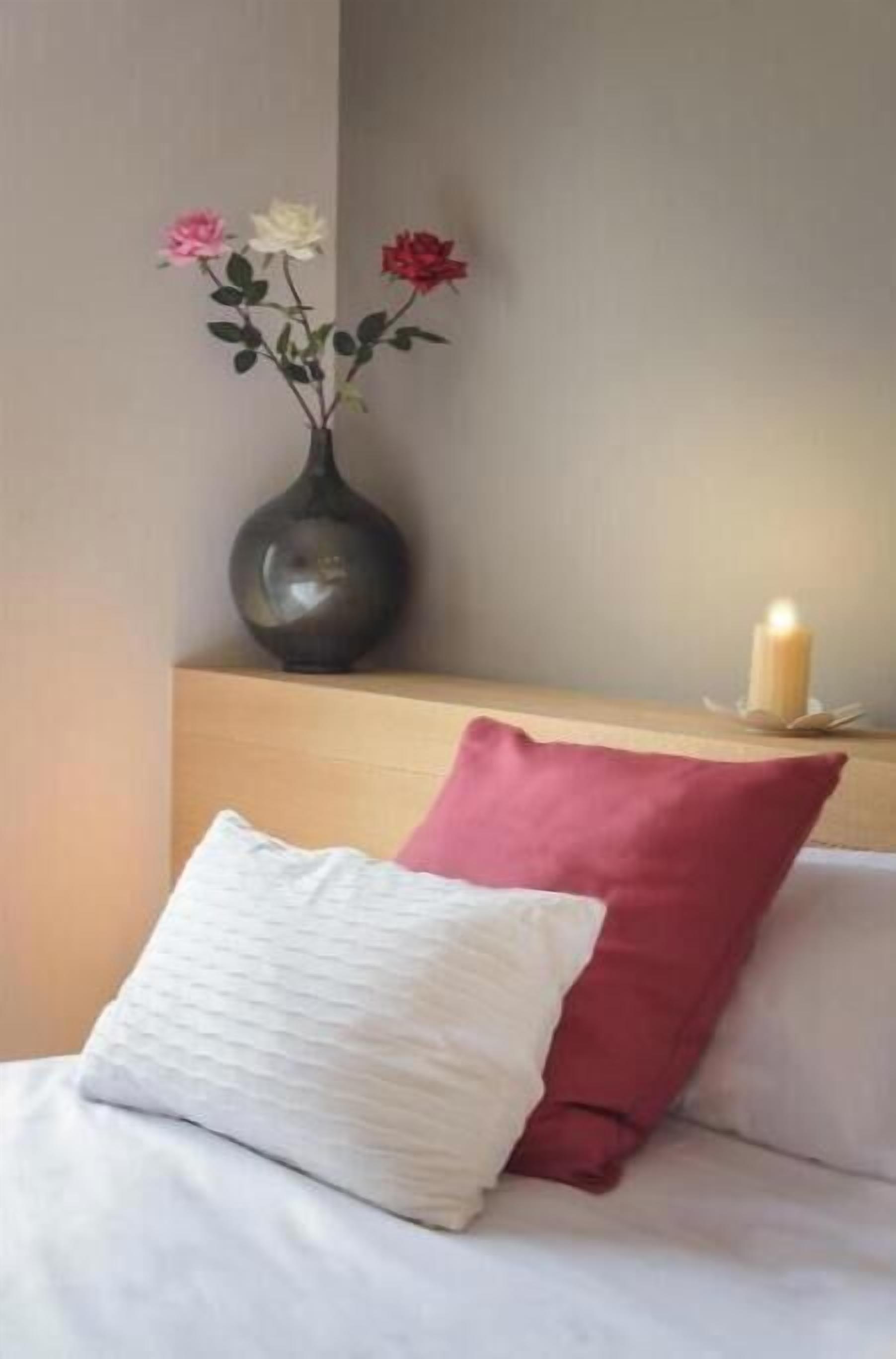 A bed with white and red pillows, a vase with flowers, and a candle on a bedside table.