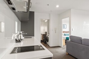 A modern kitchen with a built-in stove, a sink, and a microwave. There is a laundry area with a washing machine and a small cabinet. A grey sofa is visible in the living area. - 19th Floor 1BDR Apartment Steps from SkyCity (Auckland)