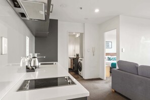 Una cocina moderna con estufa integrada, lavaplatos y microondas. Hay un área de lavandería con lavadora y un mueble pequeño. Se ve un sofá gris en la zona de estar.