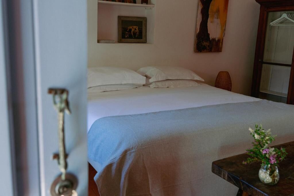 Ein ordentlich bezogenes Bett mit weißen Leinentüchern, ein kleiner Tisch mit einer Blumenvase und ein Holzschrank mit Spiegel.