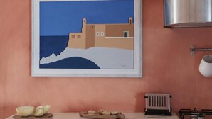 Pintura emoldurada de uma paisagem costeira com um edifício e mar, uma bancada de cozinha com uma tigela de frutas e um fogão.