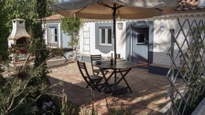 Um pátio com mesa e cadeiras, um guarda-sol e um edifício com telhado de azulejos e paredes brancas.