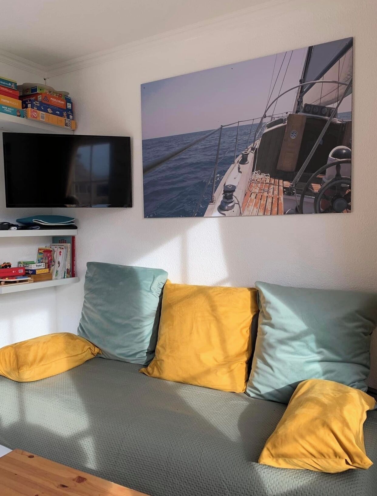 A living room with a sofa, cushions, a flat-screen TV, a bookshelf, and a painting of a sailboat on the sea.