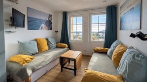 Ein gemütliches Zimmer mit einer Couch, einem kleinen Tisch und einem Fernseher. Zwei Fenster mit Vorhängen und Kunstwerken an den Wänden.