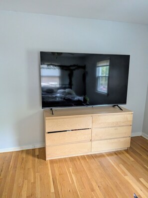 A flat-screen TV on a wooden cabinet with drawers, set against a plain wall and wooden floor.