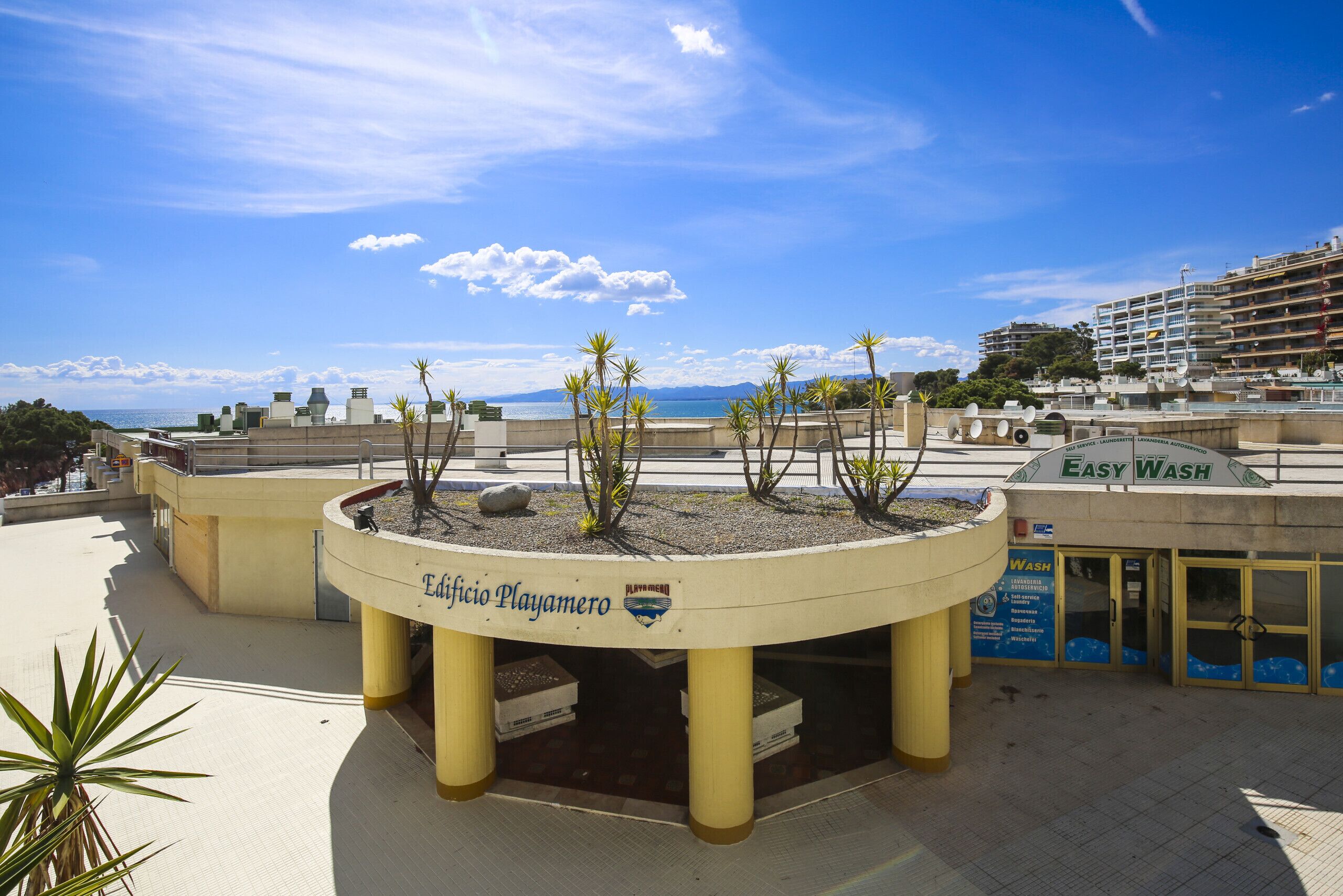 Un espacio público con un jardín circular identificado como 'Edificio Playamero', un anuncio para 'Easy Wash', y vista al mar y edificios de fondo.