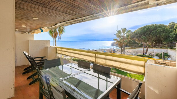 Un balcon avec une table et des chaises en verre, donnant sur une plage et une zone boisée.