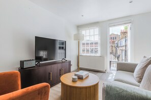 A modern living room with a flat-screen TV, a dark wooden cabinet, a round wooden coffee table, a sofa, and a lamp. - Bright Modern Flat Marylebone (London)